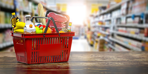Ein roter Einkaufskorb mit Lebensmitteln wie Obst, Gemüse, Brot, Milch und Fleisch steht vor unscharfen Supermarktregalen.